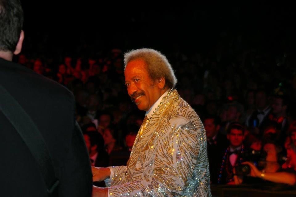 Allen Toussaint on stage in a sparkling silver-and-gold jacket, smiling back toward the band, with Chuck Credo IV in the foreground and a packed ballroom behind them.