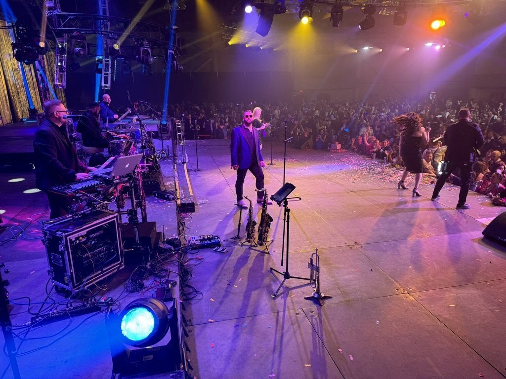 The band performing on a ballroom stage, viewed from the side of the stage, with guests dancing at the front.
