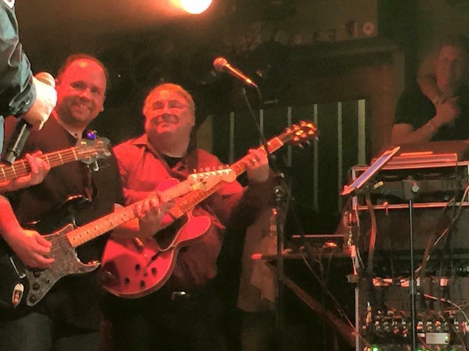 Chuck Credo IV and his father, Chuck Credo III, side by side on stage at the House of Blues, both smiling and playing guitar — a black electric and a red hollow-body.
