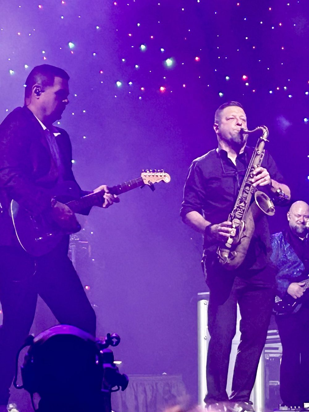 A guitarist and a saxophonist playing side by side under purple stage lights.