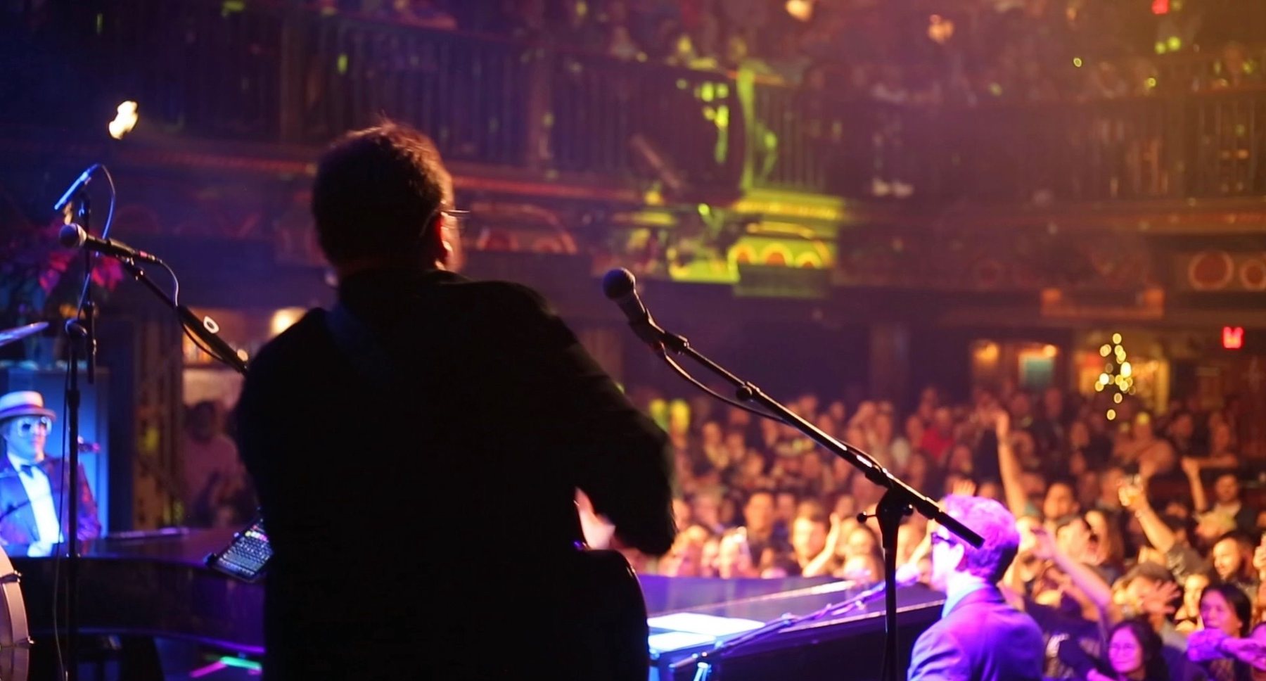 The view from the stage at a packed theatre: a guitarist seen from behind, a pianist in a boater hat and sunglasses at left, a second pianist at right, and a crowd and balconies full of raised hands.