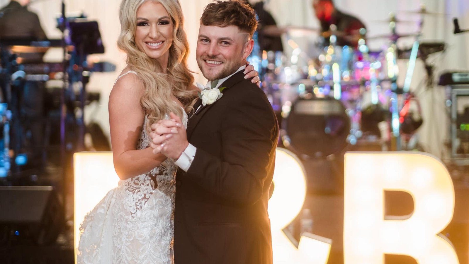 A bride and groom in their first dance in front of large lit marquee letters, the band playing behind them.