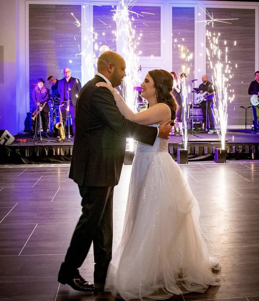 A bride and groom sharing their first dance while cold-spark fountains fire behind them and the band plays.