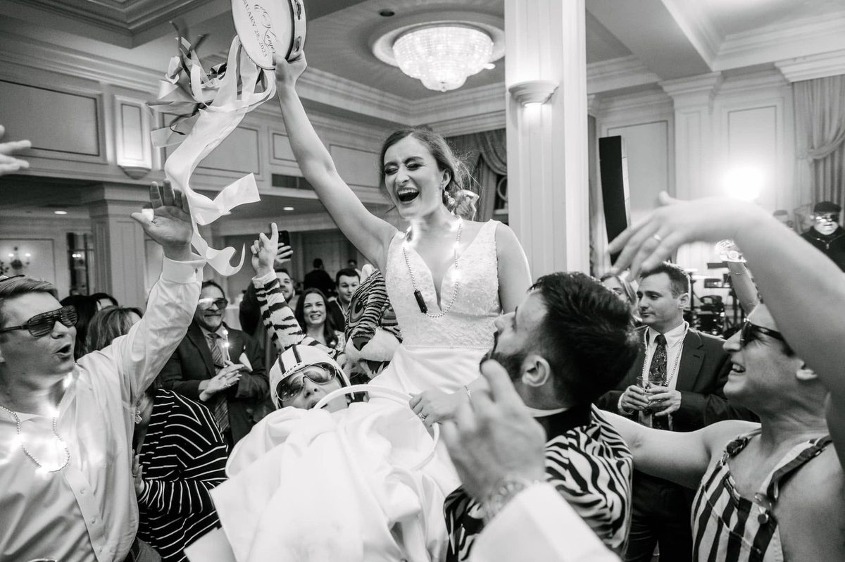 Black-and-white photo of a laughing bride lifted above the dance floor, guests cheering around her.