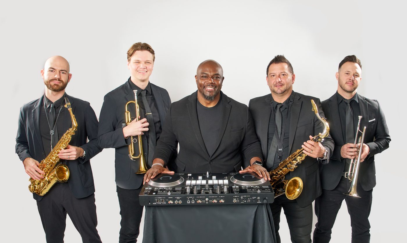 A horn section with saxophones and trumpets flanking a DJ at his decks, all dressed in black.