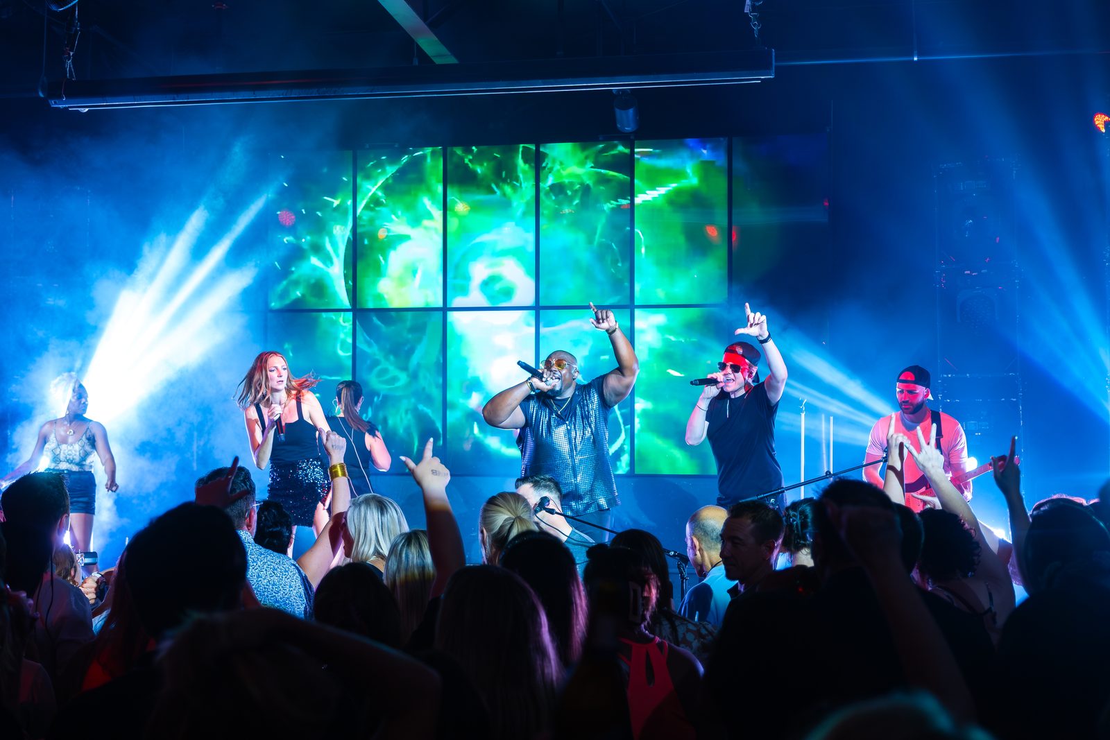 A packed crowd in front of a lit concert stage — singers out front, a giant green-lit LED wall behind them and blue beams cutting through haze.