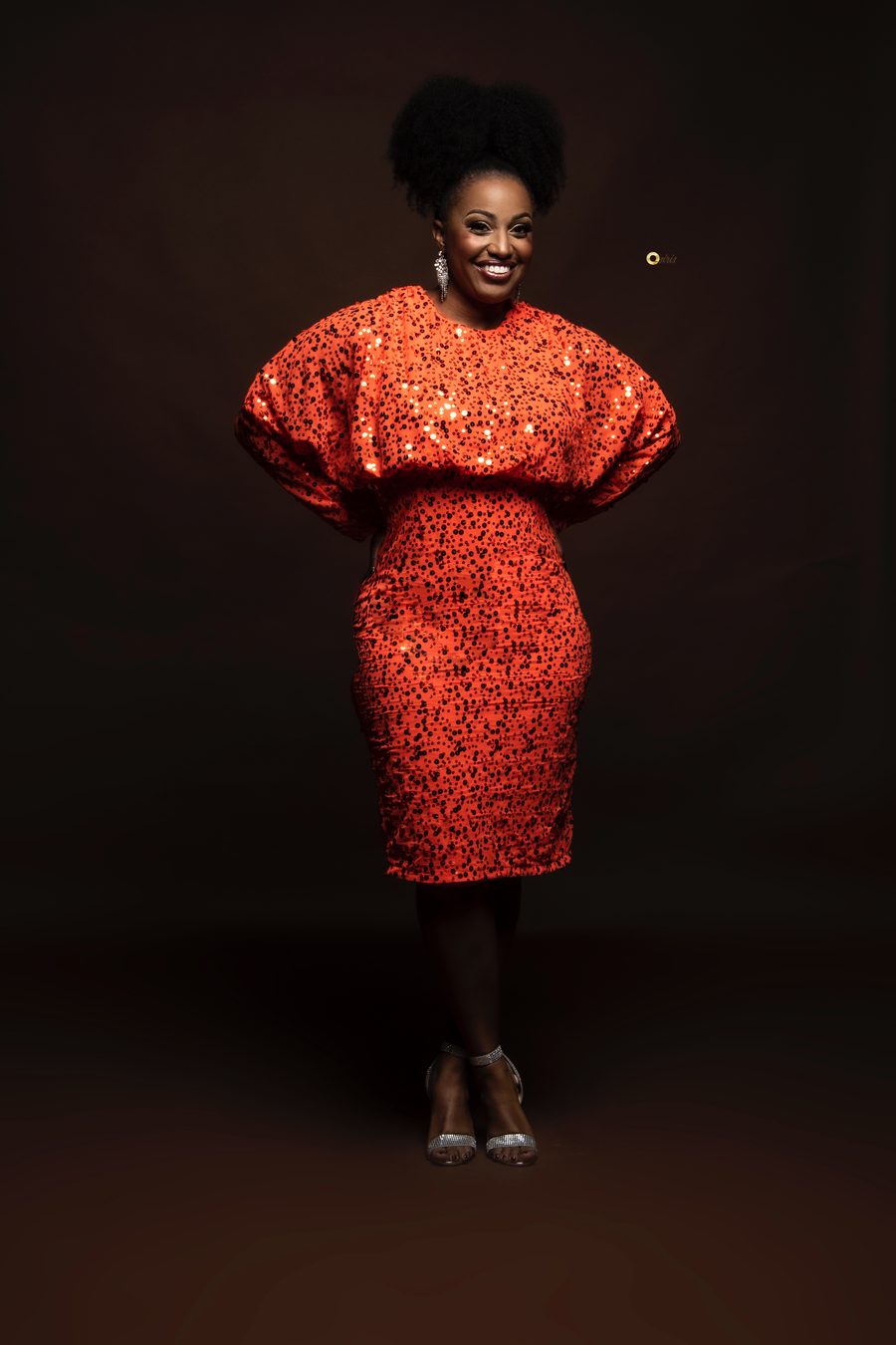 A vocalist in an orange sequined dress, smiling, against a black backdrop.